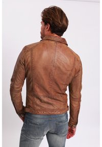 Braune Lederjacke mit tailliertem Design, sichtbaren Nähten und Reißverschlüssen; kombiniert mit hellblauen, verwaschenen Jeans.