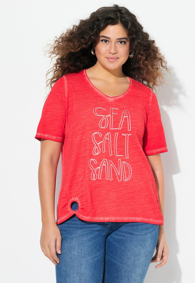 T-shirt rouge à manches courtes, arborant le texte "SEA SALT SAND" en blanc. Accent circulaire remarquable sur l'ourlet. Tissu doux et texturé.