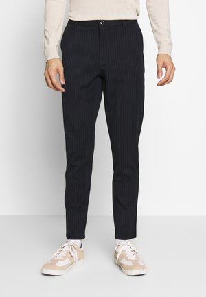 Homme portant un pantalon slim noir à fines rayures, des baskets beiges et blanches, et un haut à manches longues beige sur un fond uni.