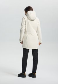 Lichtbeige waterdichte jas met een capuchon, voorzien van een getailleerd ontwerp en lange mouwen. Gecombineerd met zwarte broek en zwarte sportschoenen.