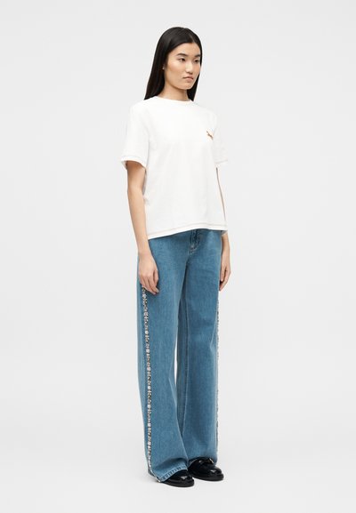 Νεαρή γυναίκα με μακριά σκούρα μαλλιά, ντυμένη με λευκό κοντομάνικο T-shirt και φαρδιά μπλε τζιν με πλευρικές ρίγες με λουλούδια, φορόντας μαύρα παπούτσια.