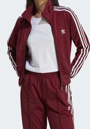 Kvinde iført en vinrød Adidas træningsdragt med hvide striber og logo, kombineret med en enkel hvid t-shirt, hænderne i lommerne.