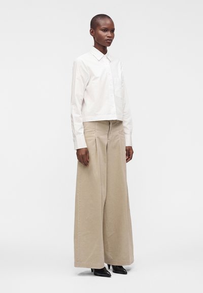 Personne portant une chemise blanche boutonnée, un pantalon beige à jambes larges et des chaussures noires à talons aiguilles pointues, debout devant un fond blanc uni.