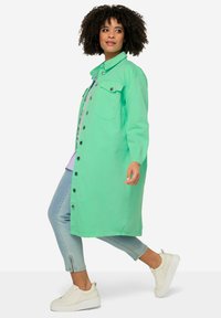 Cappotto verde con bottoni, due tasche sul petto, maniche lunghe e taglio dritto. Abbinato a jeans azzurri chiari e sneakers bianche.