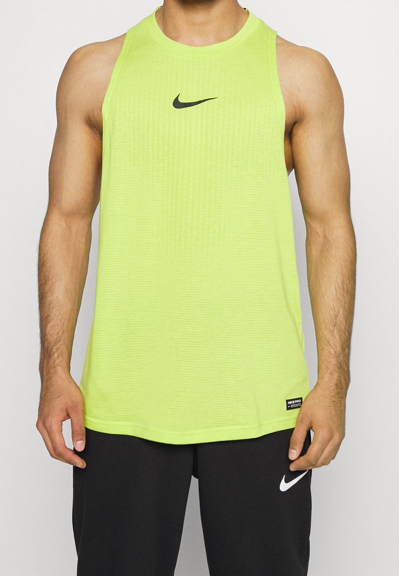 Persona musculosa vestida con una camiseta sin mangas de Nike de color lima brillante y pantalones cortos negros de Nike, de pie con los brazos relajados a los lados.