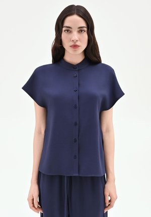 Donna con lunghi capelli scuri indossa una camicetta blu navy a maniche corte con bottoni e pantaloni abbinati, in piedi su uno sfondo bianco uniforme.