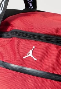 Κόκκινο σακίδιο πλάτης από υφασμάτινη ύλη με υφή, με μαύρο φερμουάρ και λευκό λογότυπο Jumpman. Μαύρο λουράκι με κείμενο ως διακόσμηση.