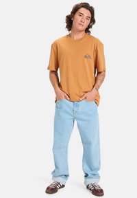 Brown cotton t-shirt with a small gray logo on the chest, paired with light blue denim jeans and brown sneakers with white accents.