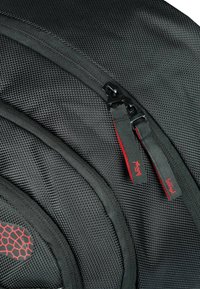 Zaino nero con tessuto testurizzato, dettagli rossi e doppie zip con marchio. Forma curva con sezioni imbottite per supporto ergonomico.