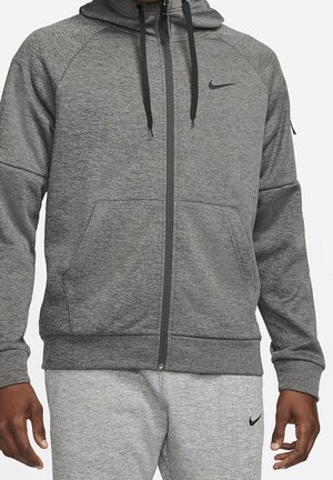 Mann trägt grauen Nike Kapuzenpullover mit Reißverschluss und schwarzen Kordeln sowie passende graue Nike Jogginghose, steht mit entspannten Armen.