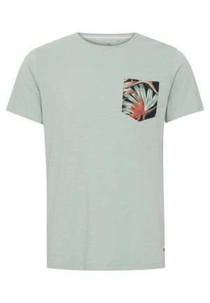 Camiseta de color verde menta claro con mangas cortas, que presenta un bolsillo negro con un estampado de hojas tropicales coloridas. Textura suave y diseño sencillo.
