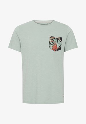 Camiseta de color verde menta claro con mangas cortas, que presenta un bolsillo negro con un estampado de hojas tropicales coloridas. Textura suave y diseño sencillo.