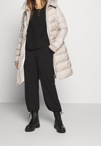 Gevuld lichtbeige jas met een capuchon, zwarte geribbelde trui, zwarte wijde broek en zwarte veterschoenen. Eenvoudig, functioneel ontwerp.