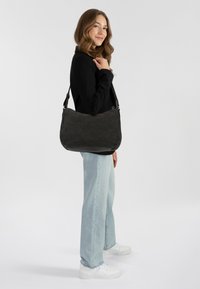 Borsa a spalla in materiale nero e morbido, con forma curvata, tracolla regolabile e hardware metallico, tenuta da una persona che indossa jeans chiari e sneakers.