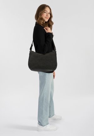 Schwarze, weiche Material Schultertasche mit gebogener Form, verstellbarem Tragegurt und metallischen Beschlägen, gehalten von einer Person in hellen Jeans und Sneakers.