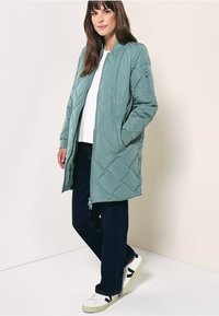 Cappotto lungo trapuntato di colore verde chiaro, con zip frontale, collo rotondo e tasche laterali, abbinato a pantaloni scuri e sneakers bianche.