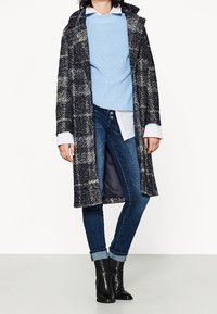 Long, navy plaid coat with white accents, textured knit blue sweater, white shirt underneath, and rolled cuff dark jeans with black ankle boots.
