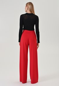 Frau von hinten zu sehen, die ein schwarzes geripptes Langarmshirt mit transparenter Rückendetails und hochtaillierte weit geschnittene rote Hose trägt.