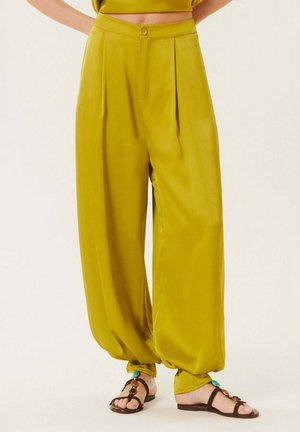 Pantalon en satin jaune moutarde taille haute, ample, avec des plis, resserré aux chevilles, porté avec des sandales plates noires ornées de perles turquoise et marron.