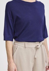 Donkerblauwe gebreide top met korte mouwen, gecombineerd met lichtbeige высокe taillebroek met een gestrikt riem en geplooide details aan de voorkant.