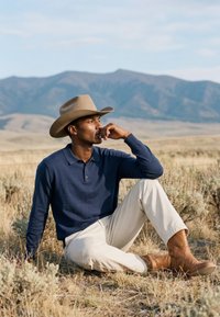 Homme portant un chapeau de cowboy, une chemise bleu marine, un pantalon crème et des bottes marron, assis sur de l'herbe sèche avec des montagnes en arrière-plan.