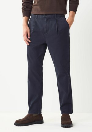 Marineblaue Hose mit Falten und seitlichen Taschen, aus einem glatten, langlebigen Stoff gefertigt. Kombiniert mit braunen Slippers und einem dunklen Pullover.