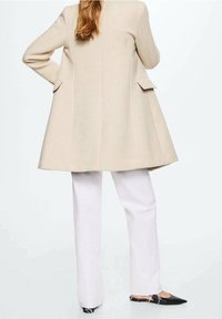 Cappotto beige su misura con silhouette svasata e tasche posteriori, abbinato a pantaloni bianchi e ballerine nere a punta. Texture liscia.