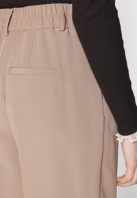 Un pantalon beige clair avec une texture lisse et une taille élastique, comportant une poche latérale et un haut noir à manches longues avec des accents en dentelle.