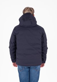 Giacca a vento blu navy con cappuccio, realizzata in un tessuto liscio e testurizzato e con una vestibilità comoda, indossata sopra jeans blu, senza accenti prominenti.