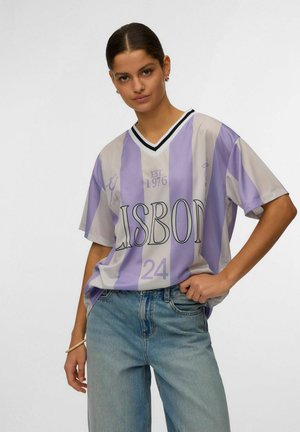 Femme portant un maillot de sport oversize à rayures violettes et blanches avec l'inscription "LISBON 24" et un jean bleu clair, posant avec une main sur la hanche.