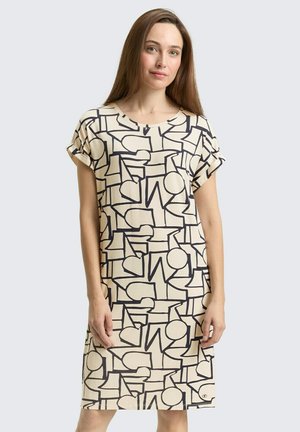 Jonge vrouw die een beige knielange jurk met zwarte abstracte geometrische patronen draagt, staand tegen een lichtgrijze achtergrond.