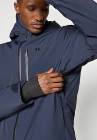 Casaco impermeável navy com capuz, fecho zip, punhos em velcro e bolsos em rede. Tecido texturizado e detalhe do logótipo no peito.