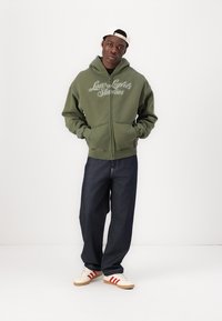 Haut zippé vert avec "Love Lights Studios" brodé sur la poitrine, associé à un jean en denim sombre ample et des baskets crème avec des rayures rouges.