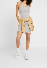 Szara bawełniana sukienka na ramiączkach z cienkimi paskami, z białym logo; zestawiona z beżową bluzą przewiązaną w talii oraz białymi sneakersami i jasnoniebieskimi skarpetkami.