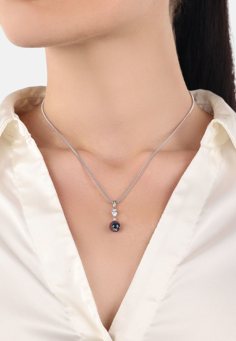 Silverfärgad kedja med en tårformad accent och en rund, mörk pärla som hänge. Bakgrunden är vit, vilket framhäver smyckets detaljer.