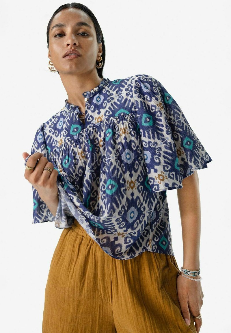 Blouse à motifs bleus et blancs avec des manches courtes évasées, un col boutonné et des fronces au niveau de la poitrine, associée à un pantalon de couleur moutarde.