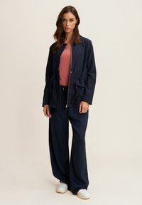Chaqueta ligera de color navy con cremallera, cintura ajustable con cordón y dos bolsillos delanteros. Lleva puesta una blusa rosa y combinada con pantalones navy a juego.