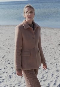 Ljust brunt ullkappa med krage, två knappar och sidofickor, tillsammans med texturerade, ribbade bruna byxor, på en sandstrand.