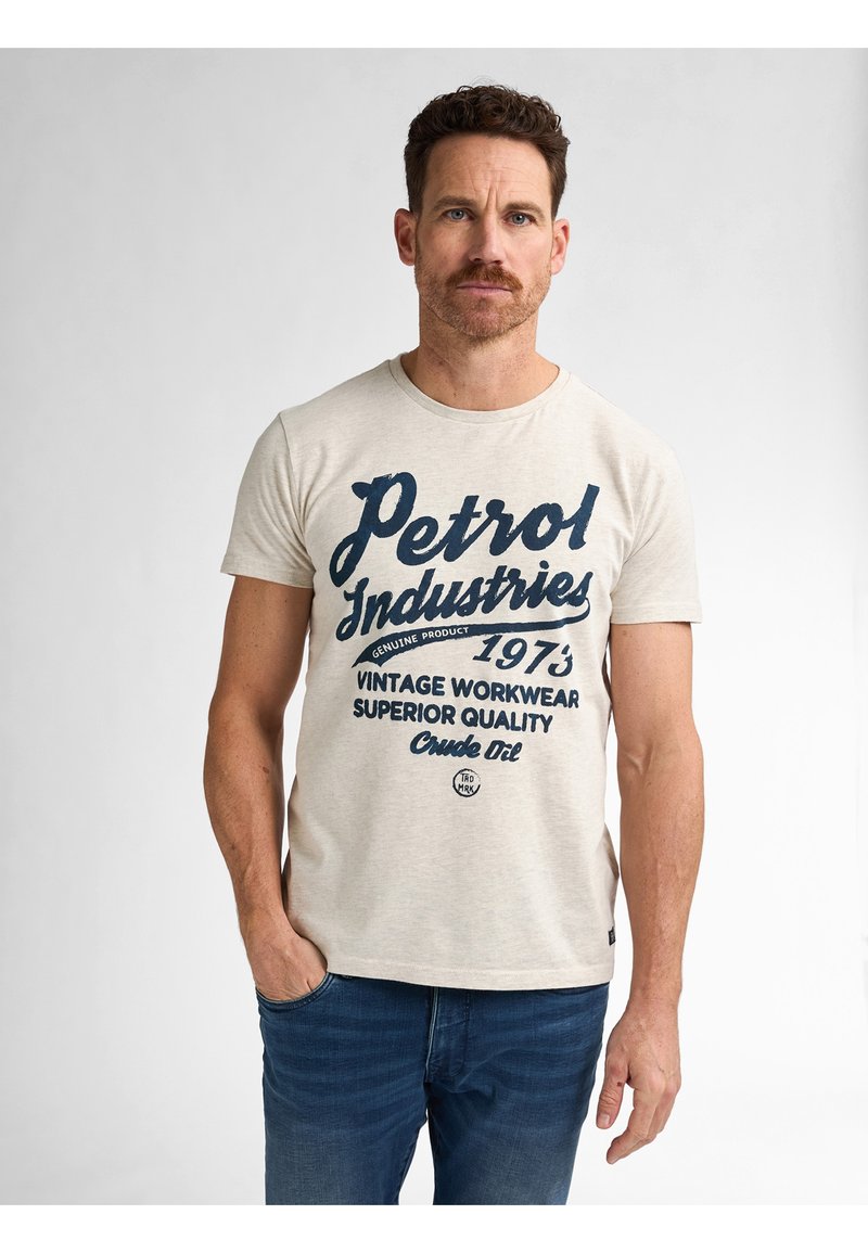Camiseta de algodón beige con un gráfico de texto y logotipo de estilo vintage en azul oscuro. Mangas cortas, cuello redondo y ajuste relajado. Se llevan jeans de denim.