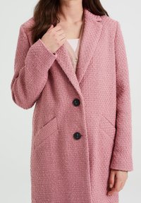 Rosa kappa i strukturerad tyg, med spetsig krage, svarta knappar och två framfickor. Medellång med en avslappnad passform.