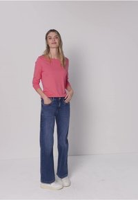 Donna in piedi contro una parete chiara e uniforme, indossa una maglietta rosa a maniche lunghe, jeans blu a gamba larga, sneakers bianche, con le mani nelle tasche e un'espressione rilassata.