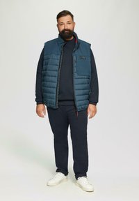 Blau gepolsterte Weste mit einem Frontreißverschluss und Seitentasche, getragen über einem marineblauen Pullover, kombiniert mit dunklen Hosen und weißen Sneakern. Einfaches Design.