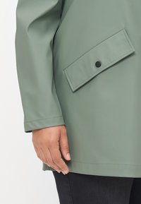 Main reposant sur le côté, portant une veste verte avec une poche à rabat fermée par un bouton noir, associée à un pantalon foncé.