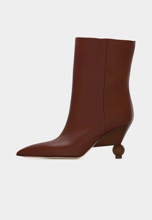 Cizmă maro din piele pentru gleznă, cu vârf ascuțit și toc rotund din lemn unic, pe un fundal simplu deschis.