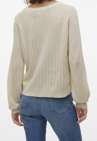 Pull en tricot beige avec des rayures ajourées verticales porté par une personne vue de dos, associé à un jean en denim bleu.