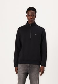 Felpa nera con zip e colletto alto, polsini e orlo a costine, con un piccolo logo sul petto. Indossata con jeans grigi chiari.