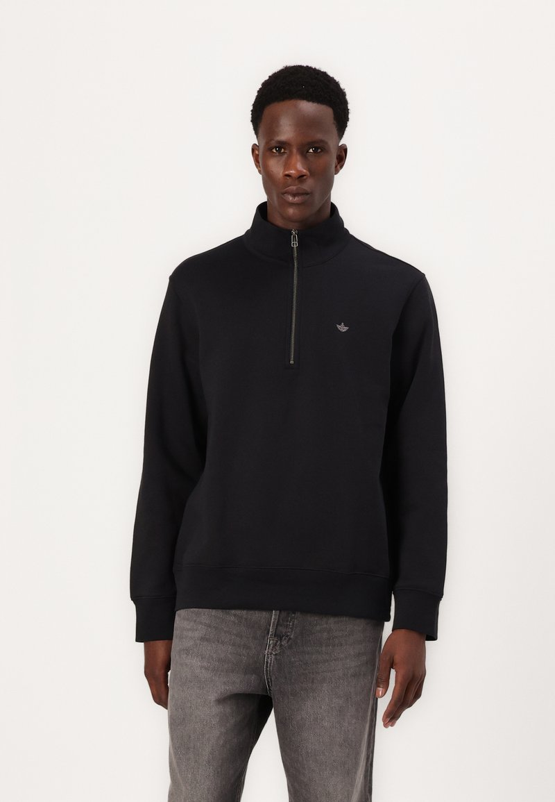 Felpa nera con zip e colletto alto, polsini e orlo a costine, con un piccolo logo sul petto. Indossata con jeans grigi chiari.