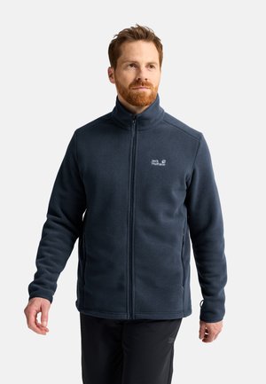 Mann mit roten Haaren, der eine marineblaue Fleecejacke mit Reißverschluss und schwarze Hose trägt, steht vor einem einfarbigen hellen Hintergrund.