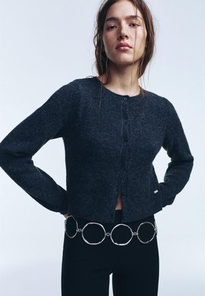 Frau trägt eine dunkelgraue, geknöpfte Strickjacke und einen schwarzen Rock mit einem großen silbernen Gürtel, der verbundene unregelmäßige runde Ringe aufweist.