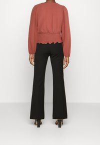 Blusa di colore ruggine, con texture, maniche a sbuffo e orlo scallop, abbinata a pantaloni neri svasati e tacchi aperti.